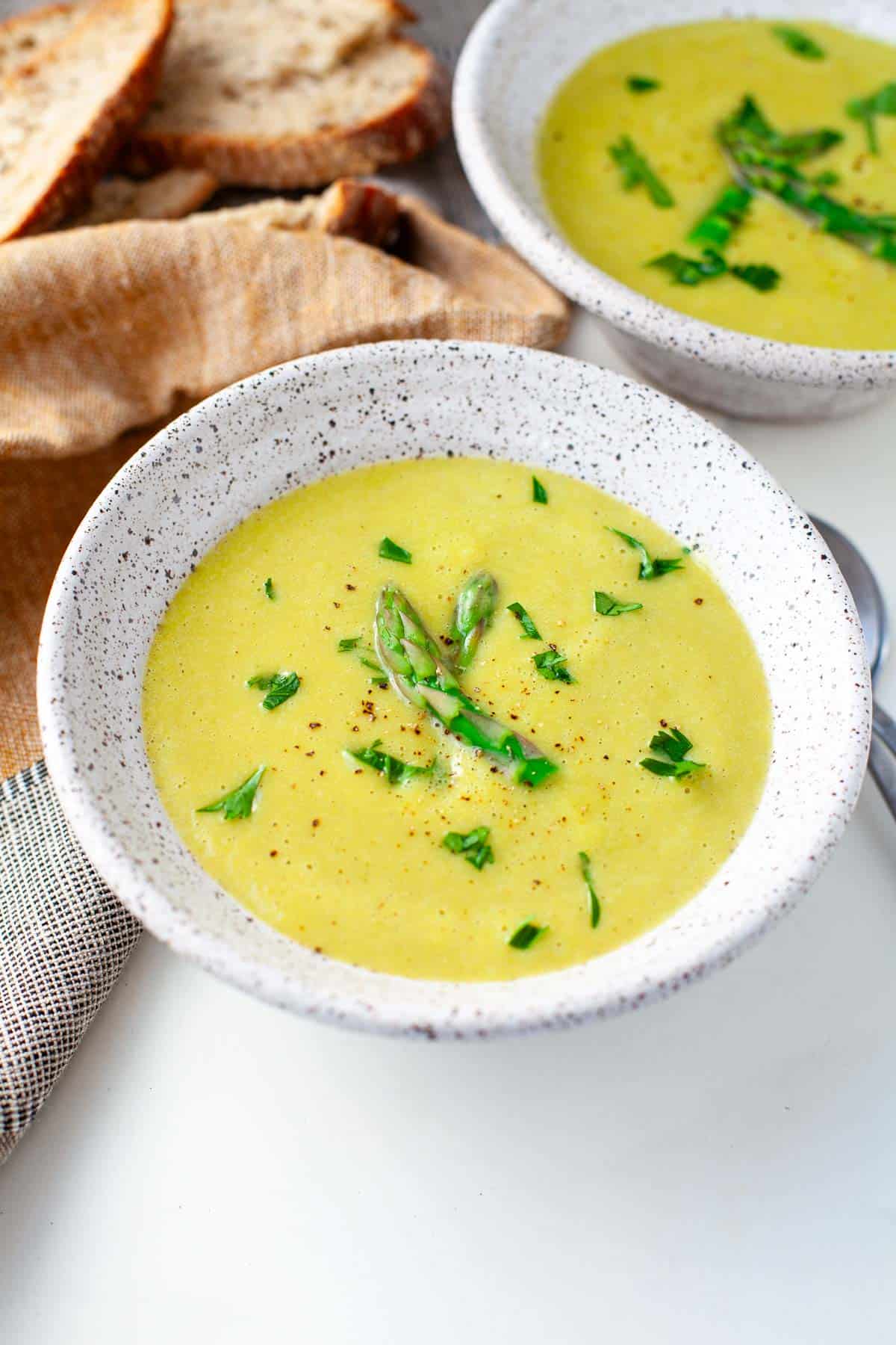 two white speckled bowls of vegan asparagus soup garnished with chopped asparagus and parsley beside a kitchen towel